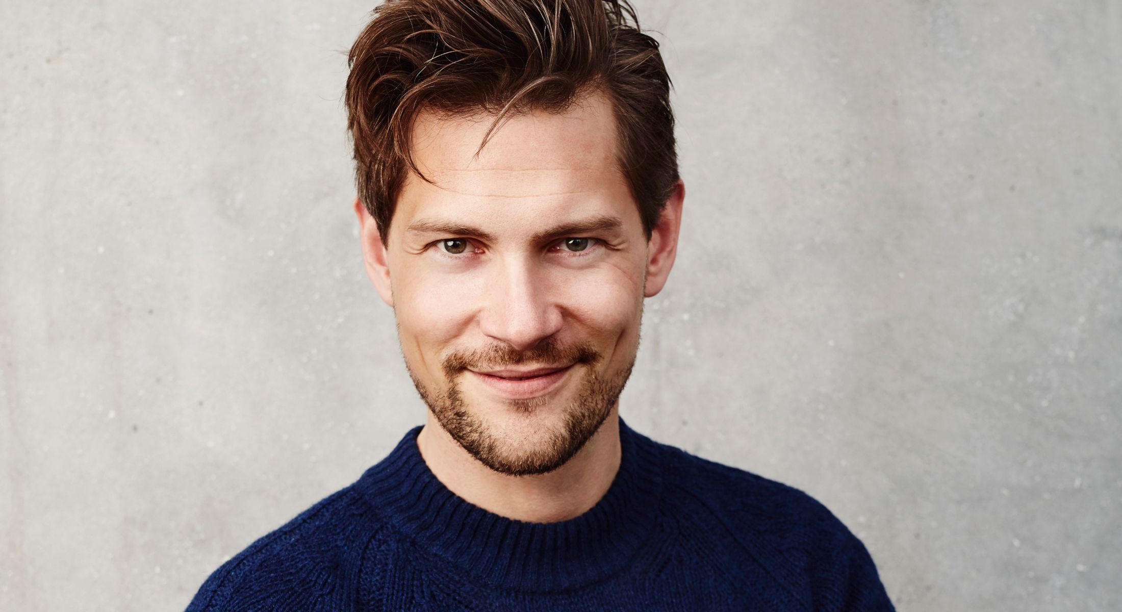 Smiling man in a blue sweater against concrete background.