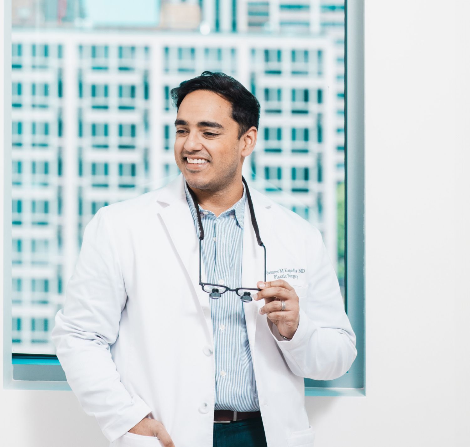 Chicago plastic surgeon, Dr. Sameer Kapadia, holding glasses in modern office.